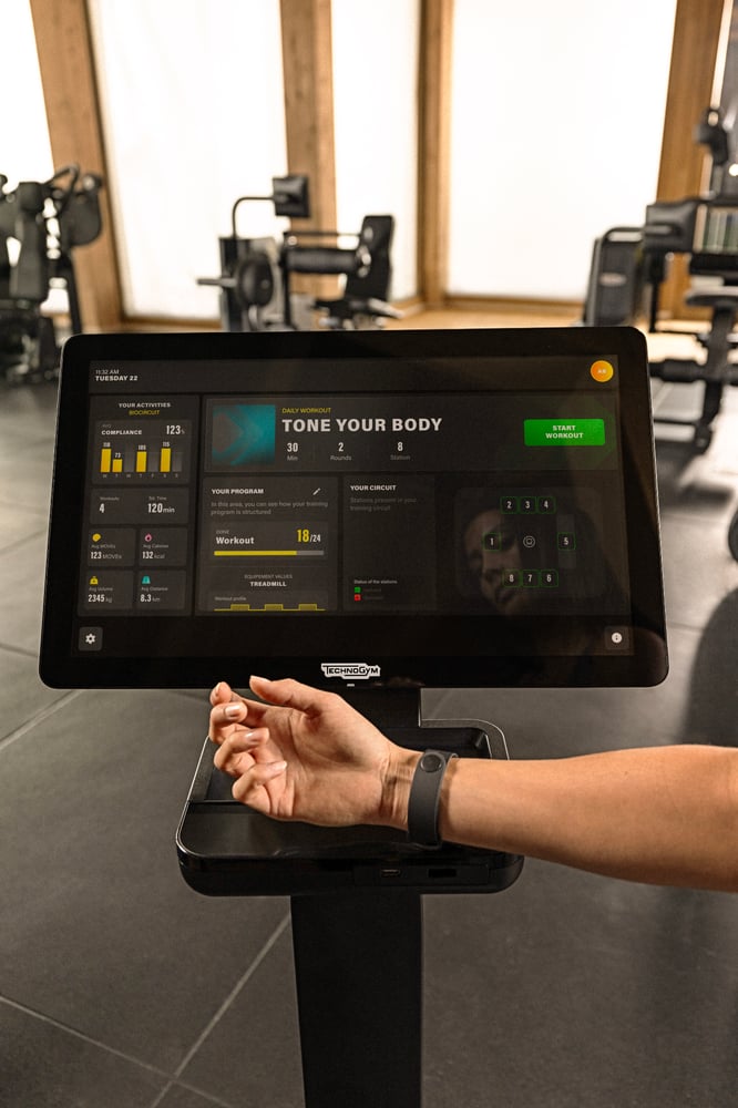 Käsi älykello ranteessa Technogym-laitteen näytön edessä, jossa näkyy treeniohjelma.